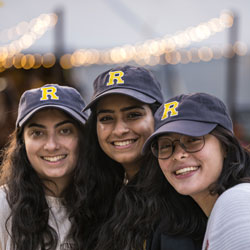 A group of smiling young adults sits at a table, each wearing white sunglasses and matching blue hats. They are surrounded by gift boxes, pamphlets, and floral arrangements. The setting appears to be a casual event or gathering.